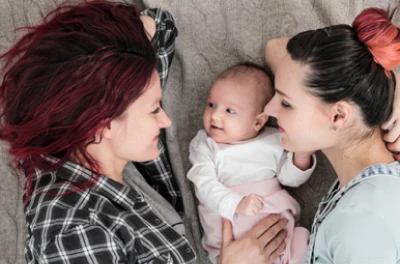 Two adults lie on either side of a baby, smiling and making eye contact. The family shares a warm, affectionate moment on a soft blanket.
