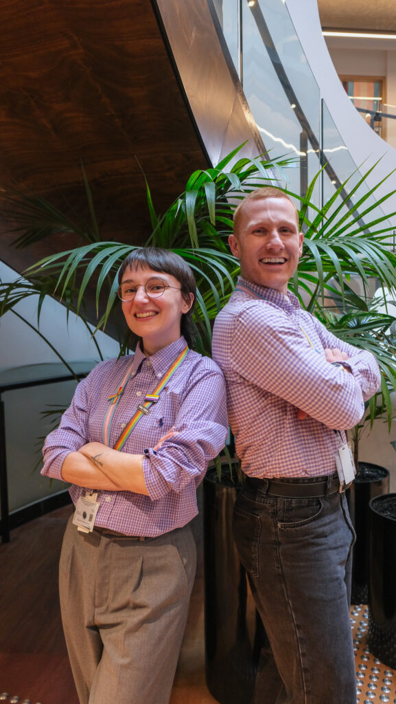 Two smiling people wearing rainbow pins and lanyards standing back-to-back indoors, representing LGBTQIA+ workplace inclusion and community support.