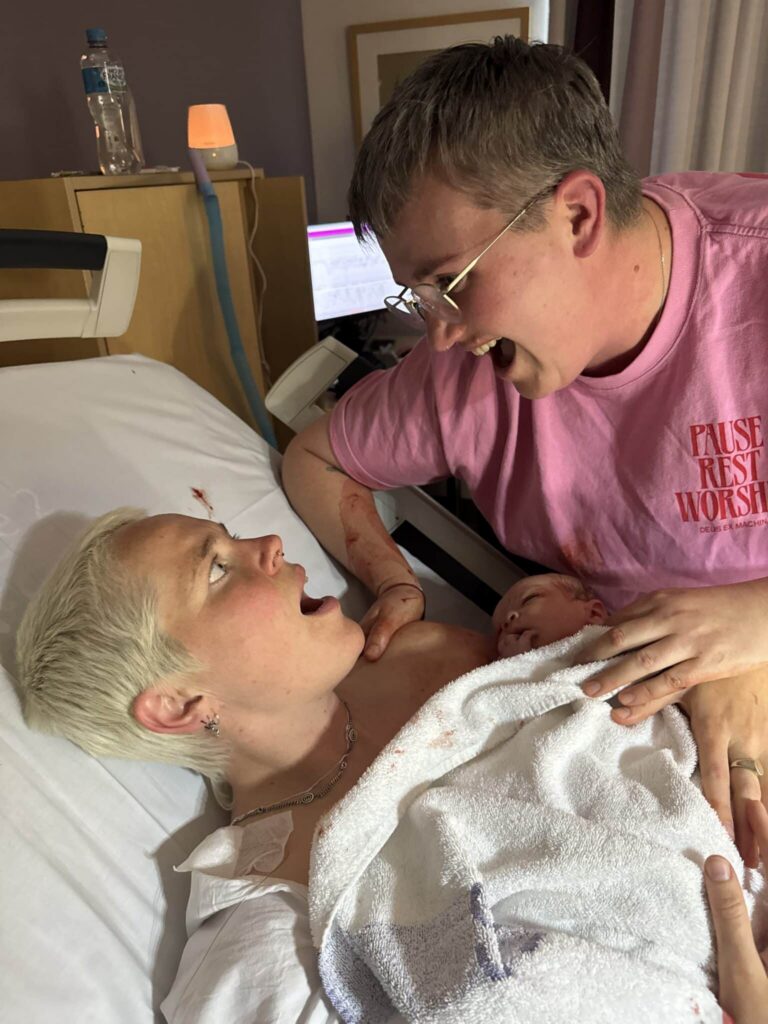 A newborn rests on one parent's chest as the other leans in smiling. Both gaze at their baby with joy in a hospital room moments after birth.