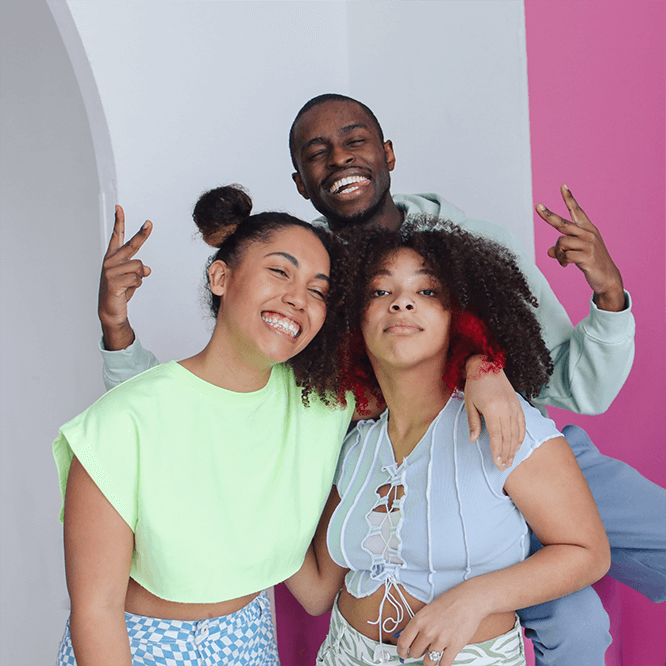 Three young people smiling and posing together with their arms around each other in a colourful indoor space.