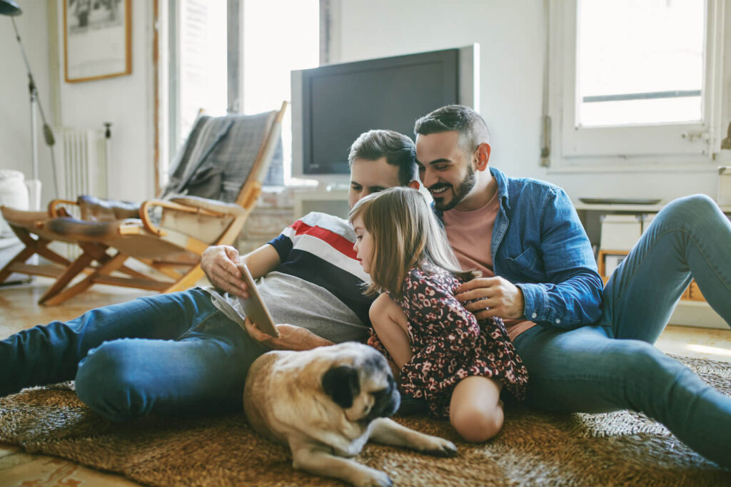 Two parents and a young child sitting together on the floor at home, looking at a tablet, with a dog lying beside them.