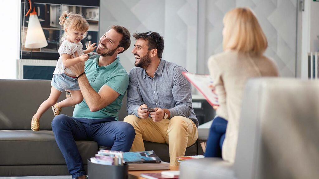 Two men on a sofa smile as a little girl hugs one of them; a woman sits opposite with a clipboard in a bright waiting area.
