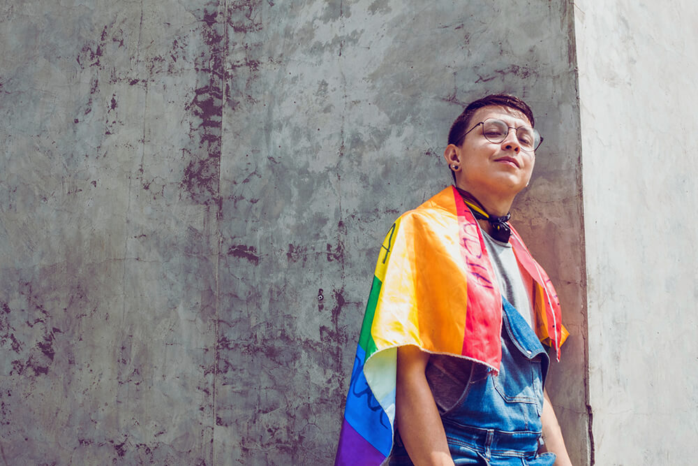 A person wearing a rainbow Pride flag over their shoulders, standing confidently against a concrete wall.