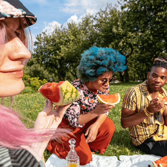 Three young people enjoying watermelon together during a picnic in a park on a sunny day.