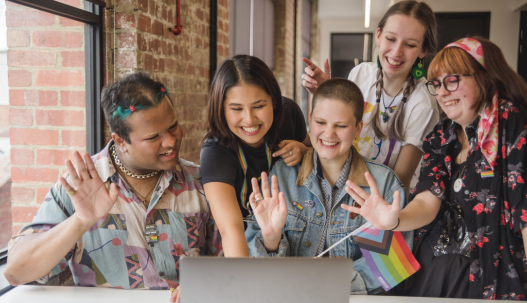 Five smiling LGBTQIA+ young people gather around a laptop, waving during a video call while holding a Progress Pride flag in a bright community space.