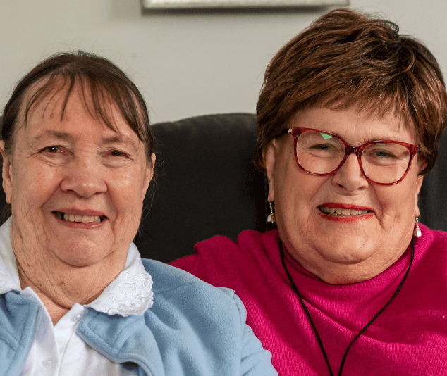 Two older women sit side by side, smiling warmly at the camera, sharing a close and affectionate moment together indoors.