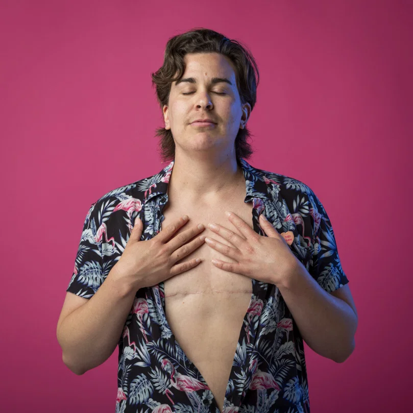 Steff Fenton stands against a pink background, wearing an open floral shirt and showing their chest.