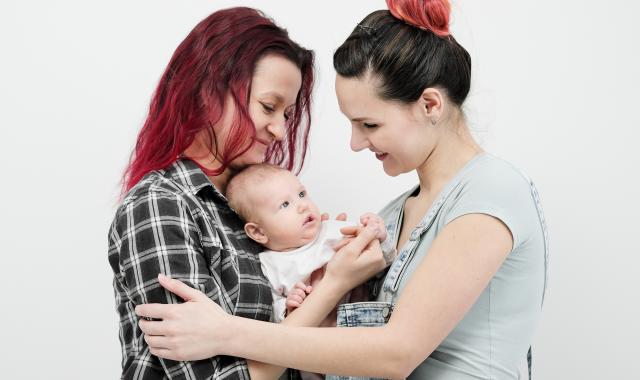Two mothers smile at their baby while holding hands around them, sharing a warm and loving family moment against a white background.