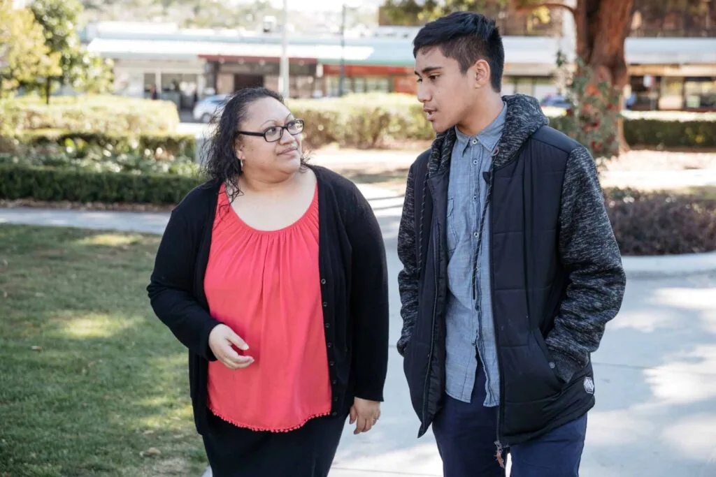 An adult and teenager walk side by side in a park, talking together. The adult listens attentively as the young person speaks.