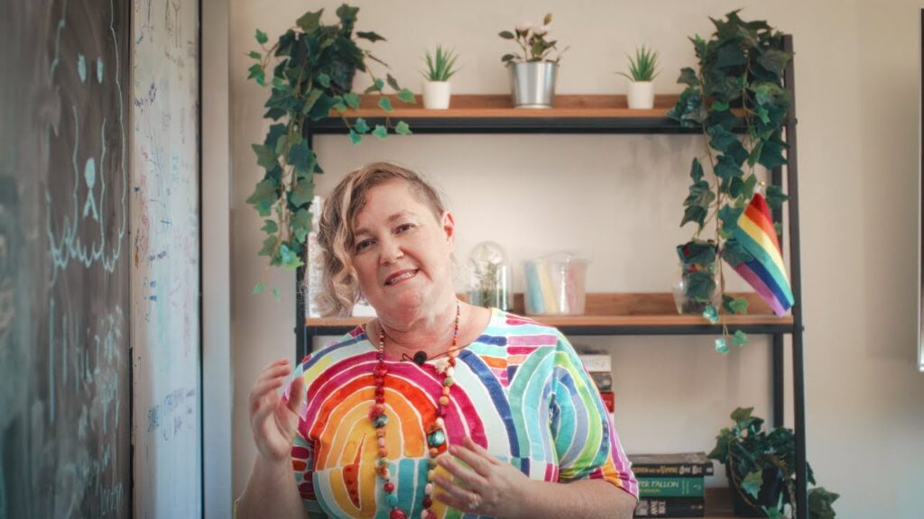 Michelle Jeffries stands in a bright room beside a whiteboard, speaking with hands raised. Michelle wears a colourful rainbow-patterned top and beaded necklace. There are plants and a small Pride flag on shelves behind Michelle.