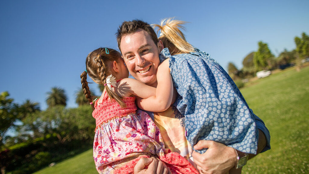 Parent embraces two young children outdoors in a park, celebrating family connection and belonging.