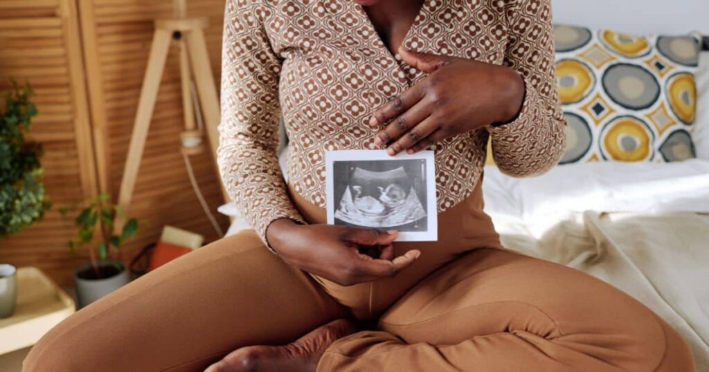 A pregnant person sitting on a bed holding an ultrasound scan in front of their stomach in a warm, home setting.