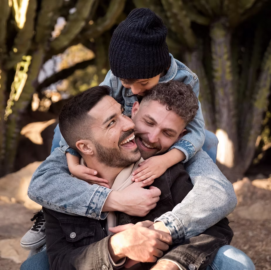 Two parents hugging a young child outdoors, smiling and laughing together.